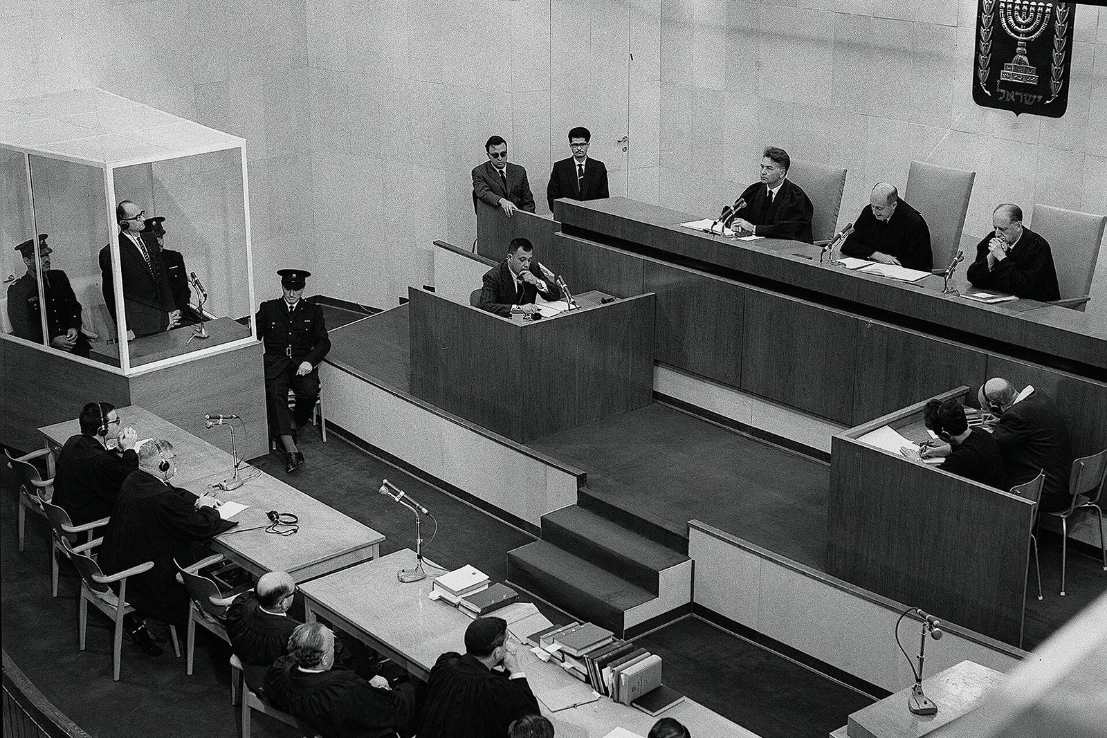 Black-and-white view down into the Jerusalem courtroom, with the defendant in a glass booth at left and the judges on the bench at right.